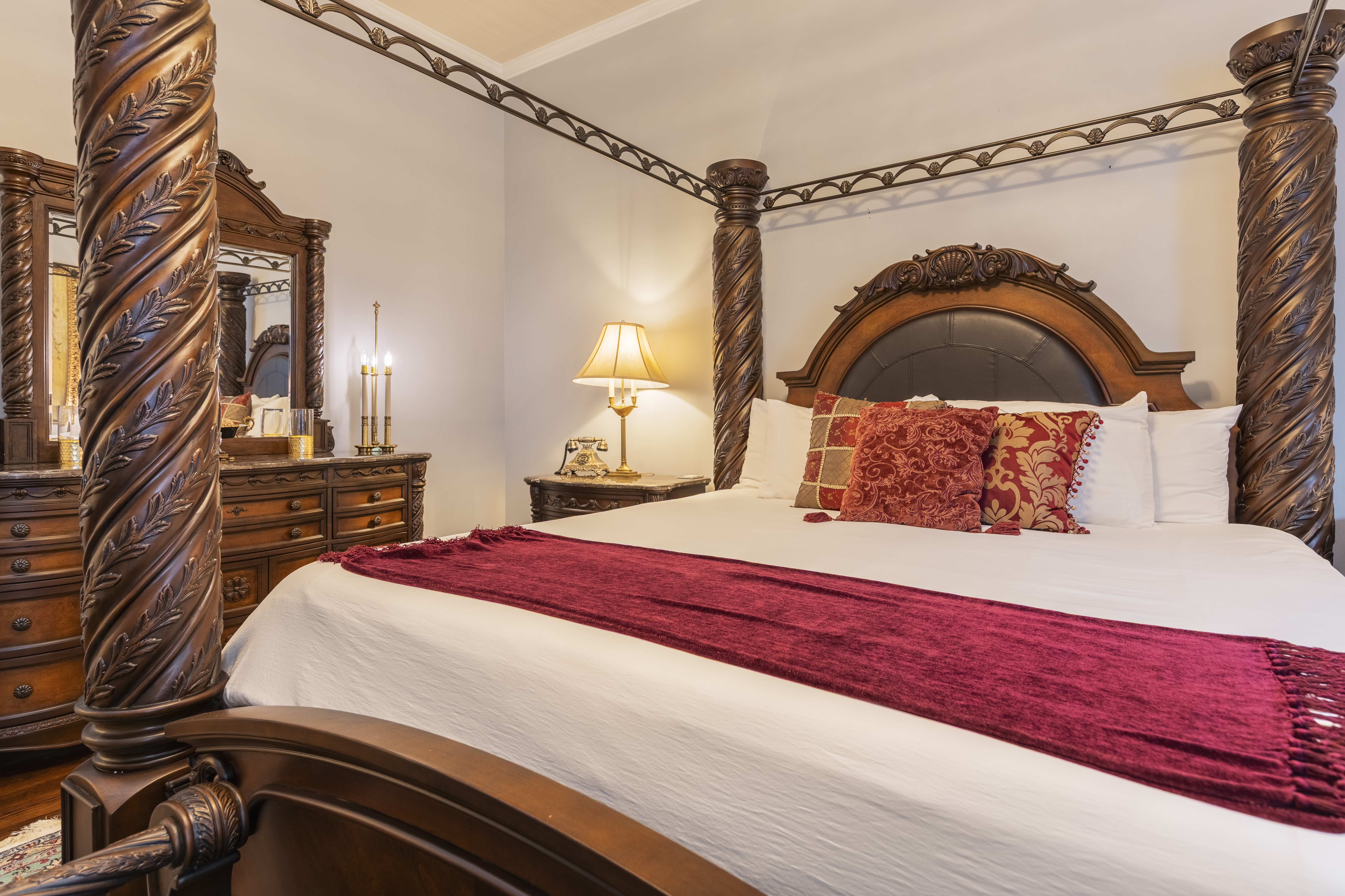 Elegant bedroom with a large, ornate canopy bed, rich red accents, and carved wooden pillars. A warm lamp and dresser create a cozy, luxurious feel.