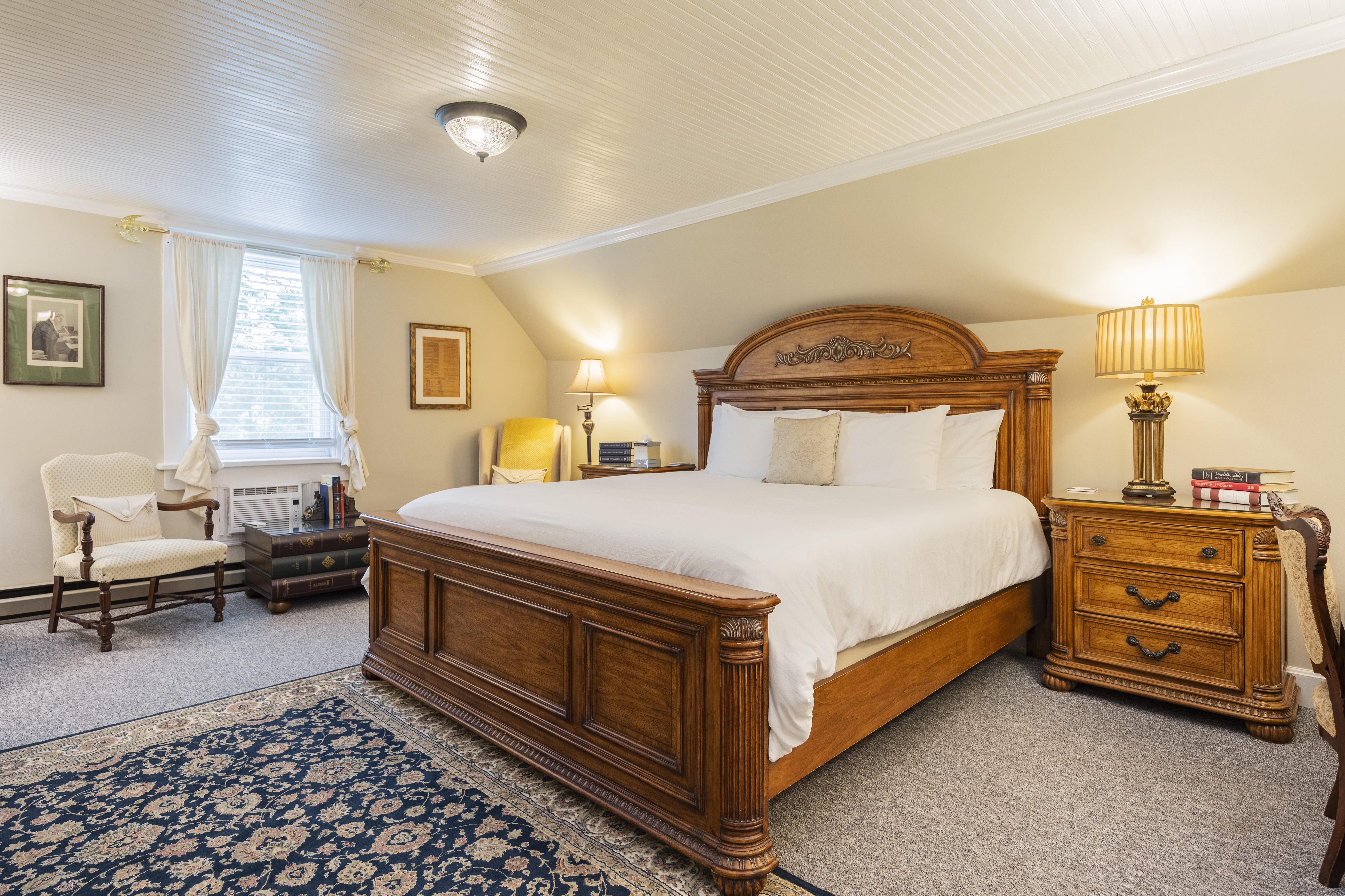 Cozy bedroom with a large wooden bed, white bedding, and soft lighting from two table lamps. Elegant armchairs and a window with curtains add warmth.