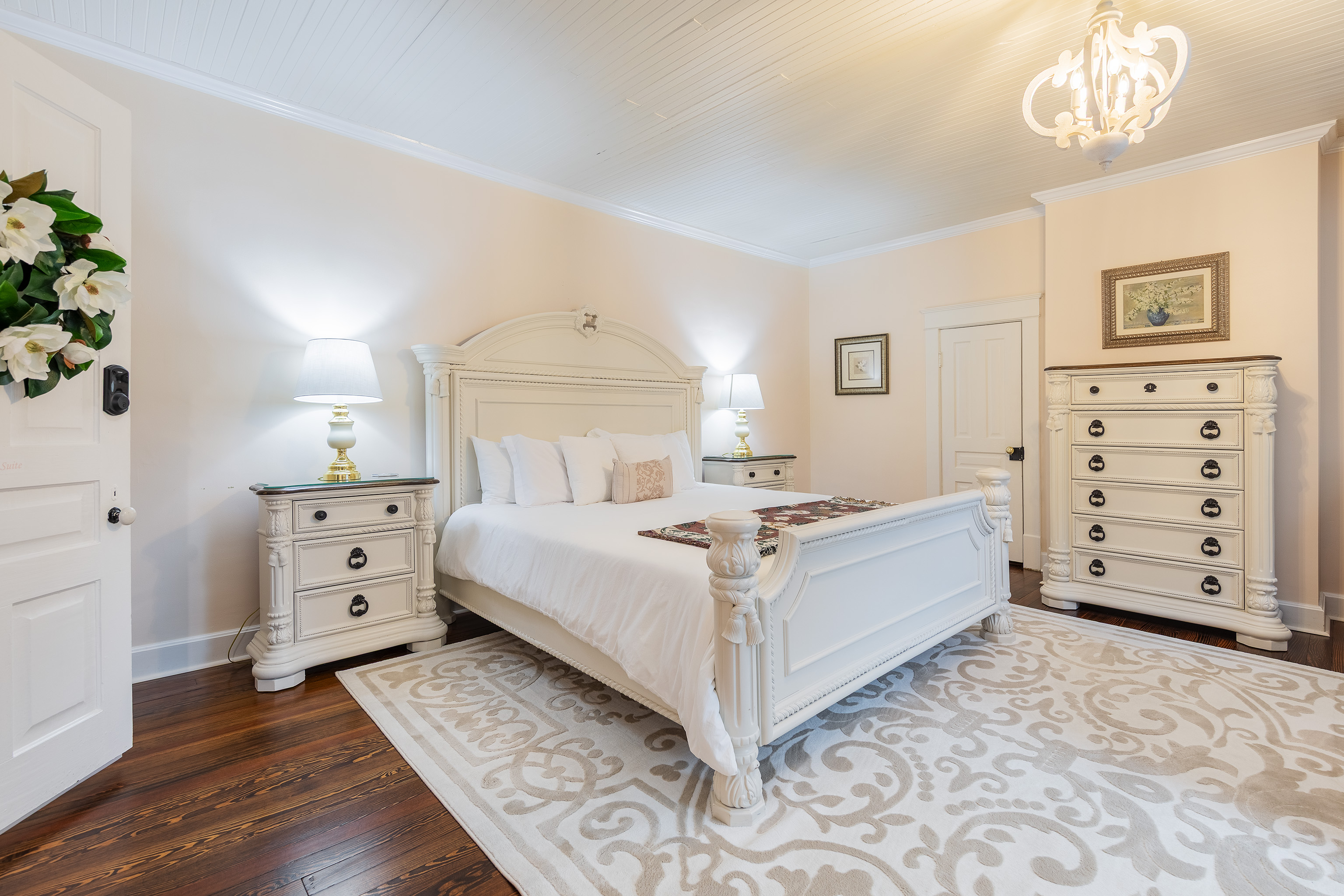 Cozy Magnolia Suite bedroom with a white antique bed, matching nightstands, and lamps. The room features hardwood floors, a decorative rug, and a magnolia wreath on the door.