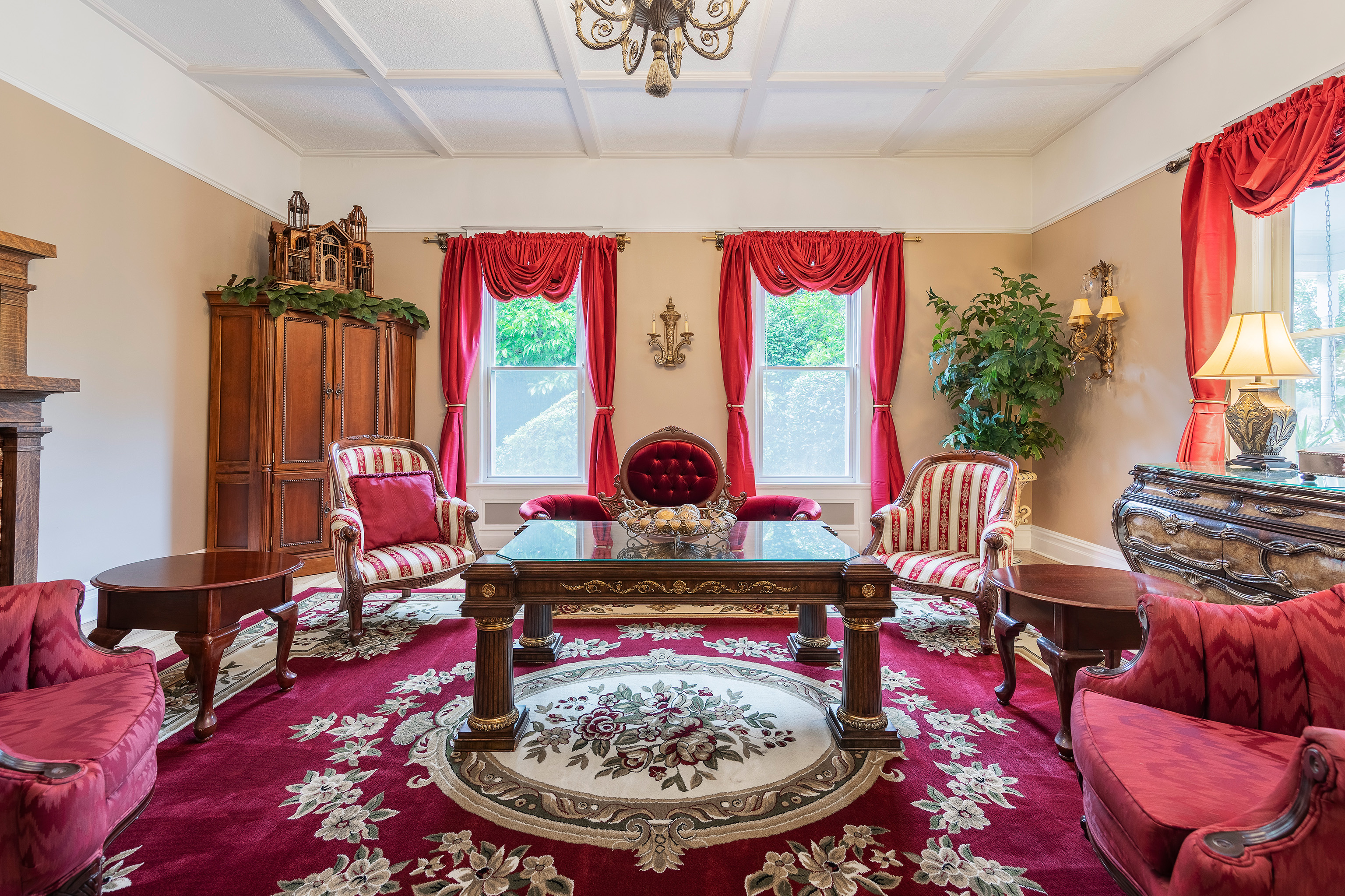 Elegant living room with vintage decor, showcasing ornate wood furniture, red floral carpet, and matching drapes. Sunlight streams through two windows.