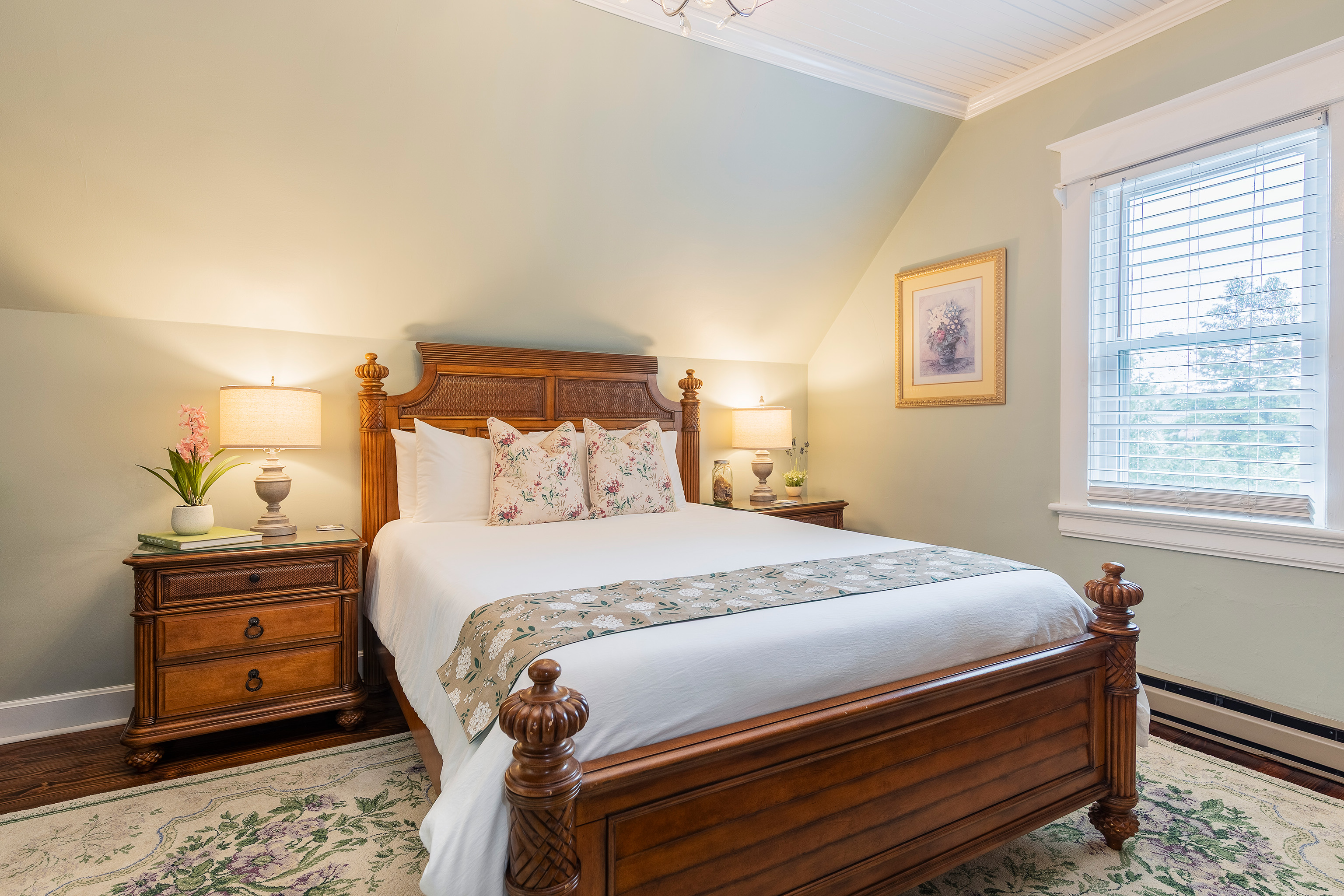 Cozy bedroom with a wooden bed, floral bedding, and soft lighting from lamps on wooden nightstands. A framed picture hangs on a light green wall.