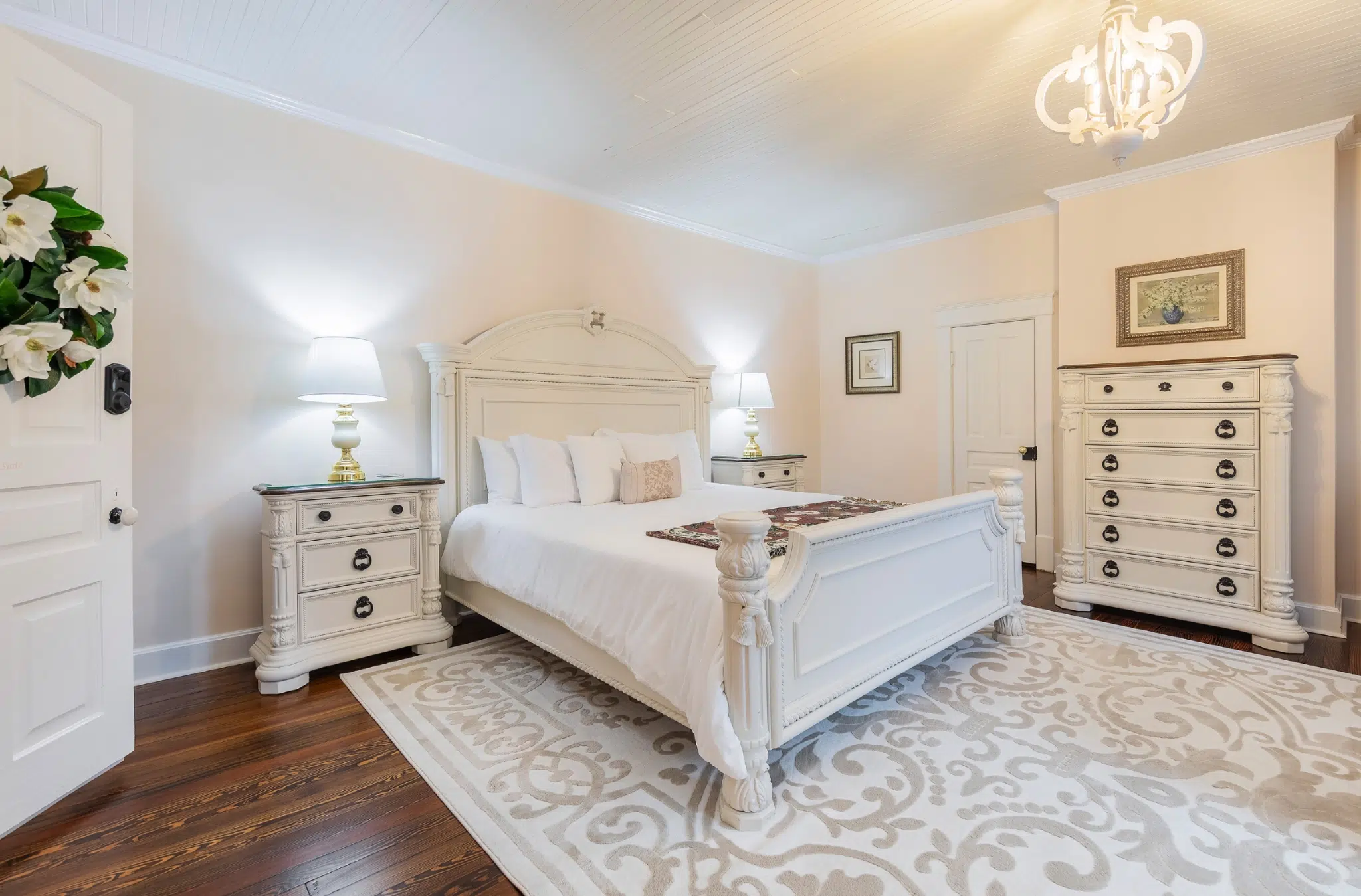 Elegant bedroom with a large white bed, ornate bedside tables with lamps, and a matching dresser. Soft beige rug on wooden floor, and floral door wreath. Cozy ambiance.
