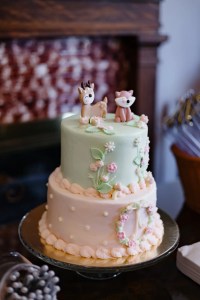 Two-tier pastel cake with deer and fox toppers. Decorated with pink flowers and leaf accents, creating a whimsical, woodland theme.