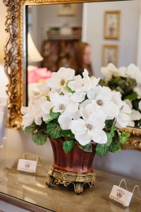 Elegant bouquet of large white flowers in a decorative vase sits on a table. A gilded mirror reflects soft interior details, creating a warm ambiance.