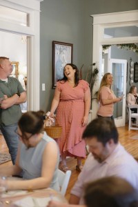 A woman in a pink dress stands smiling with a wicker basket at a social gathering. Guests in casual attire chat and laugh in a warmly lit room.