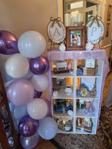 A decorative shelf adorned with purple and white balloons. Framed photo, books, and elegant ornaments on the shelves exude a celebratory ambiance.