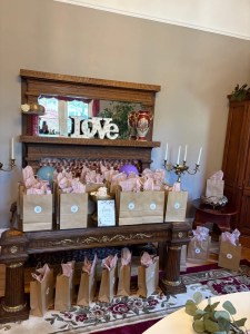 Gift bags with pink tissue paper are arranged on a decorative wooden table under a mirror. The word 