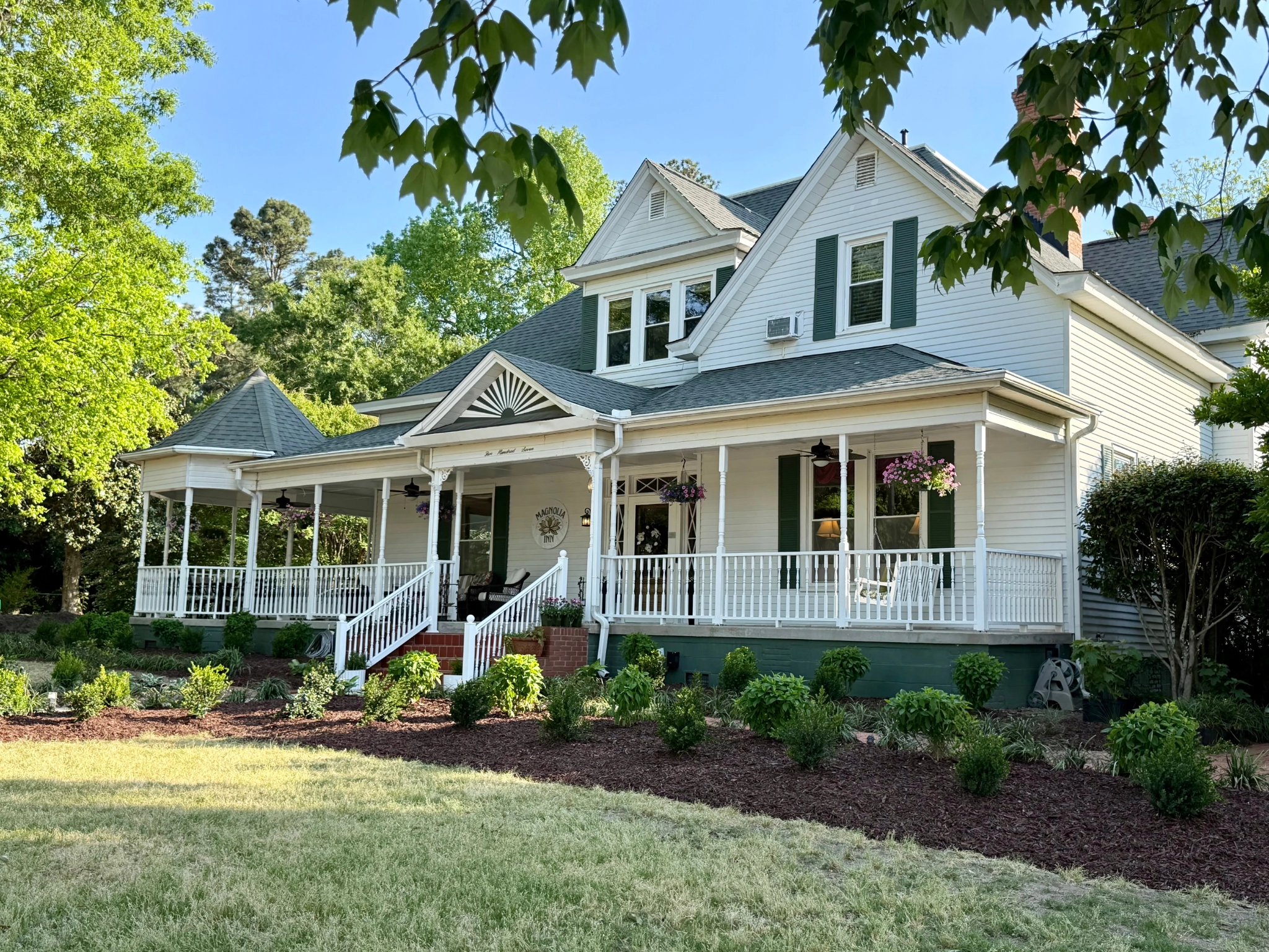 Victorian-style house with a large porch, white railings, hanging flower baskets, and lush garden. Sunlit and inviting, exuding charm and elegance.