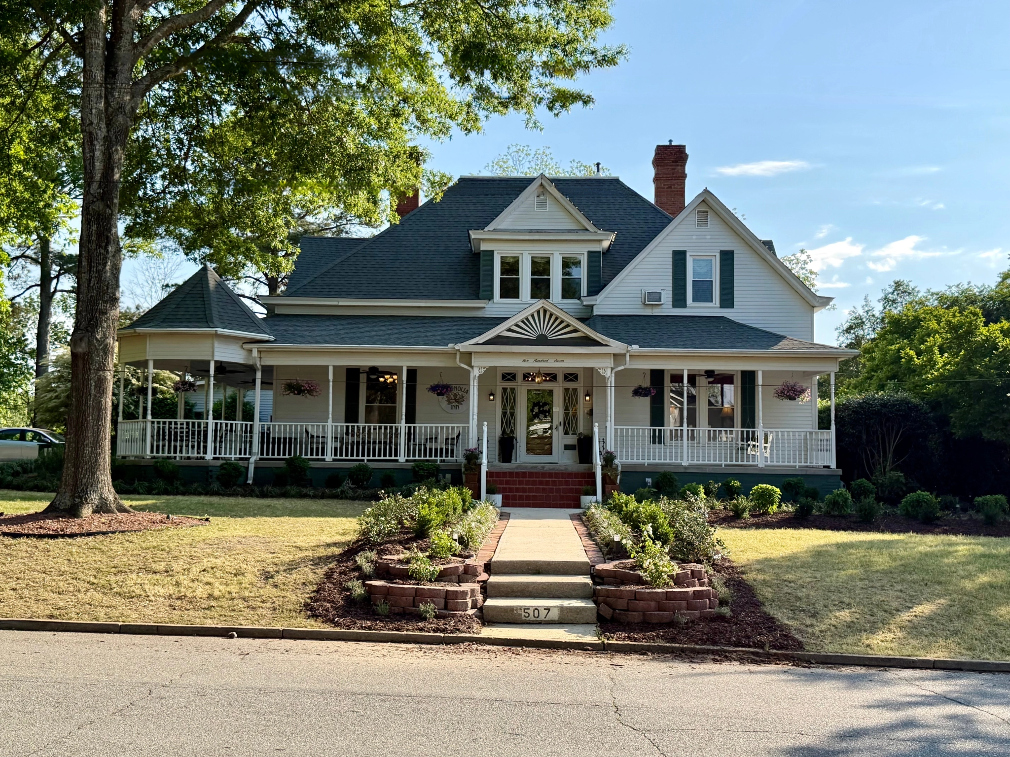 Victorian-style house with a wraparound porch, white siding, and dark roof, set amidst lush greenery. A welcoming path leads to the front door.