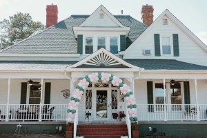 A charming white house with green shutters features a festive balloon arch in pastel colors over the entrance, conveying a welcoming and celebratory tone.