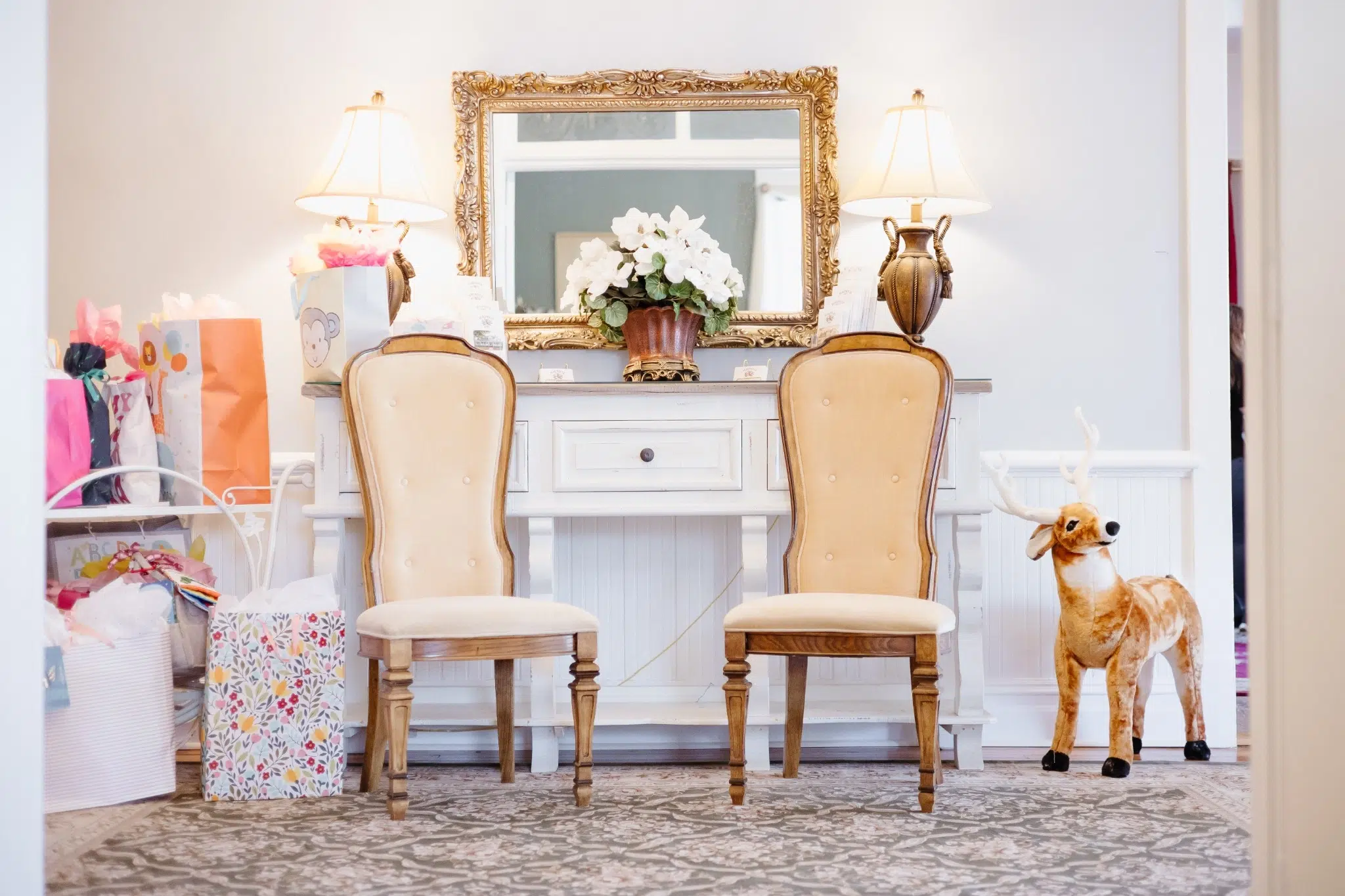 Elegant room with two vintage chairs, a white console table, ornate mirror, lamps, and potted flowers. Gift bags and a plush reindeer on the side.
