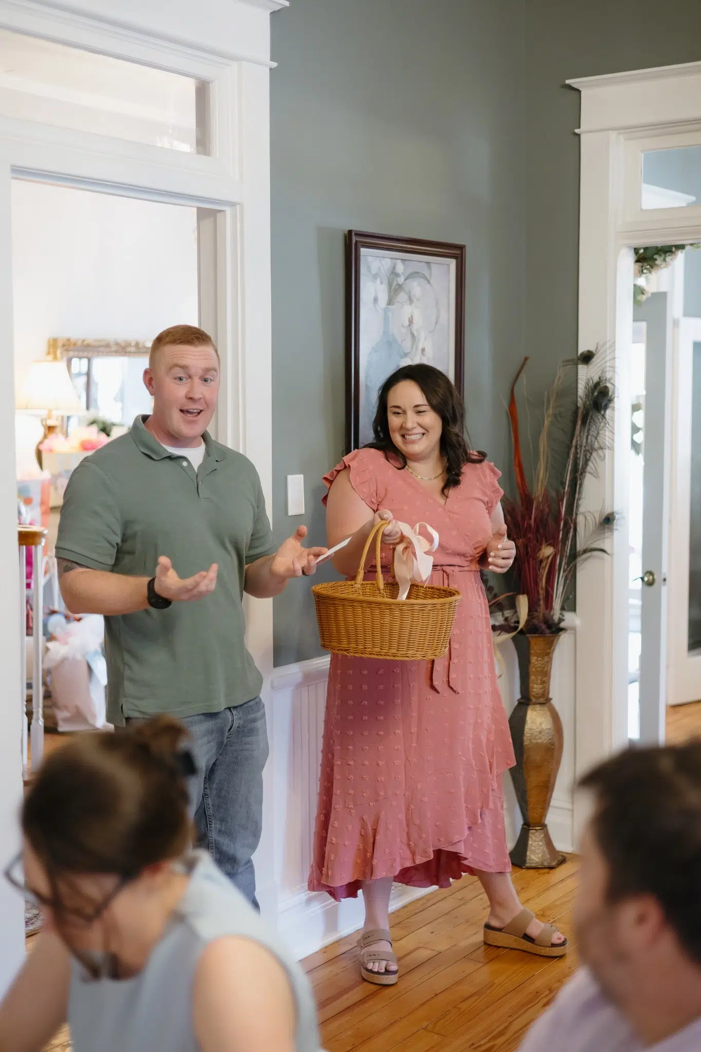 A man and woman in casual attire stand in a living room, smiling warmly. The woman holds a wicker basket. The atmosphere is joyful and welcoming.