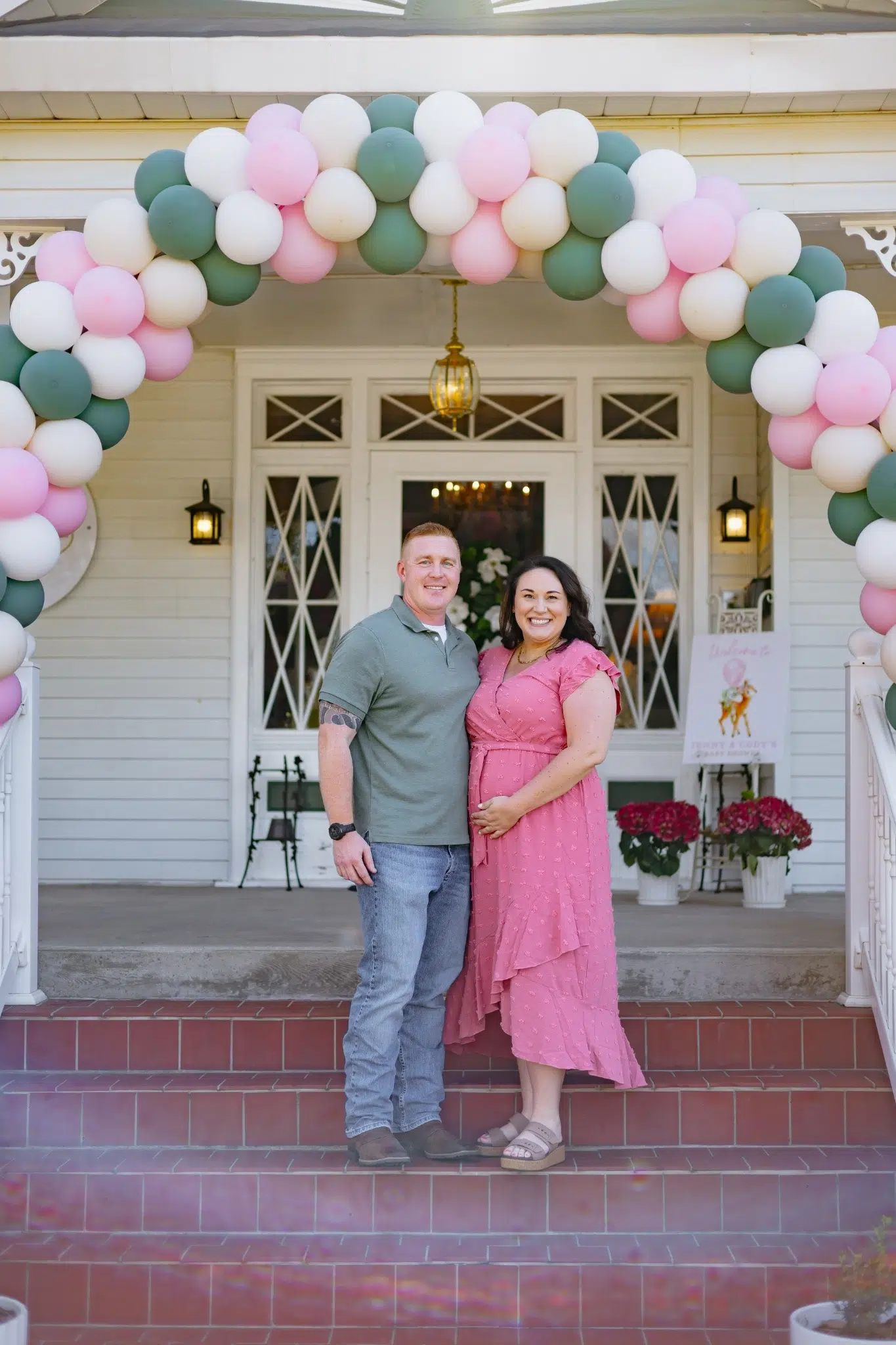 A smiling couple stands under a pink, green, and white balloon arch on a porch. The woman cradles her belly, suggesting a joyful, celebratory event.