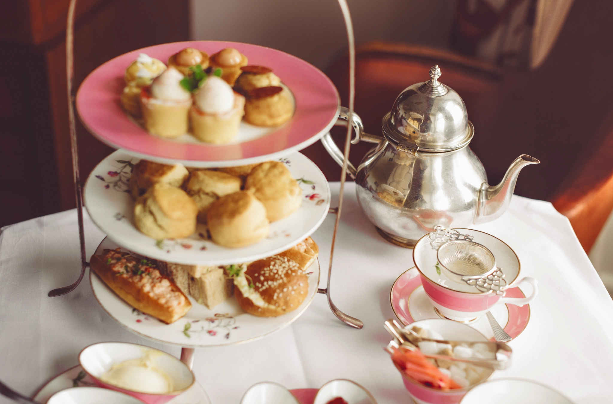 A three-tiered stand with assorted pastries, scones, and sandwiches next to a silver teapot and pink teacup set on a white tablecloth, evoking elegance.
