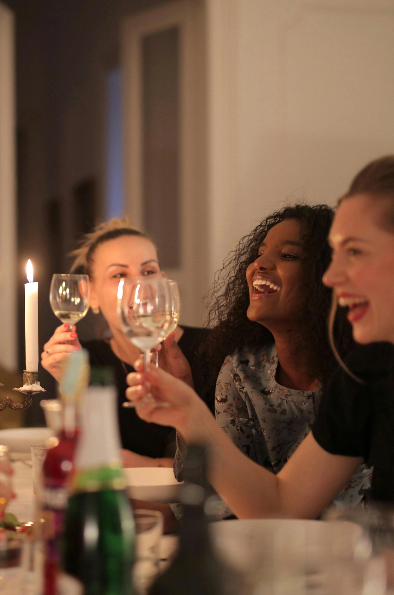 Three women joyfully toast with wine glasses at a candlelit dinner. The atmosphere is warm and lively, conveying a sense of celebration and friendship.