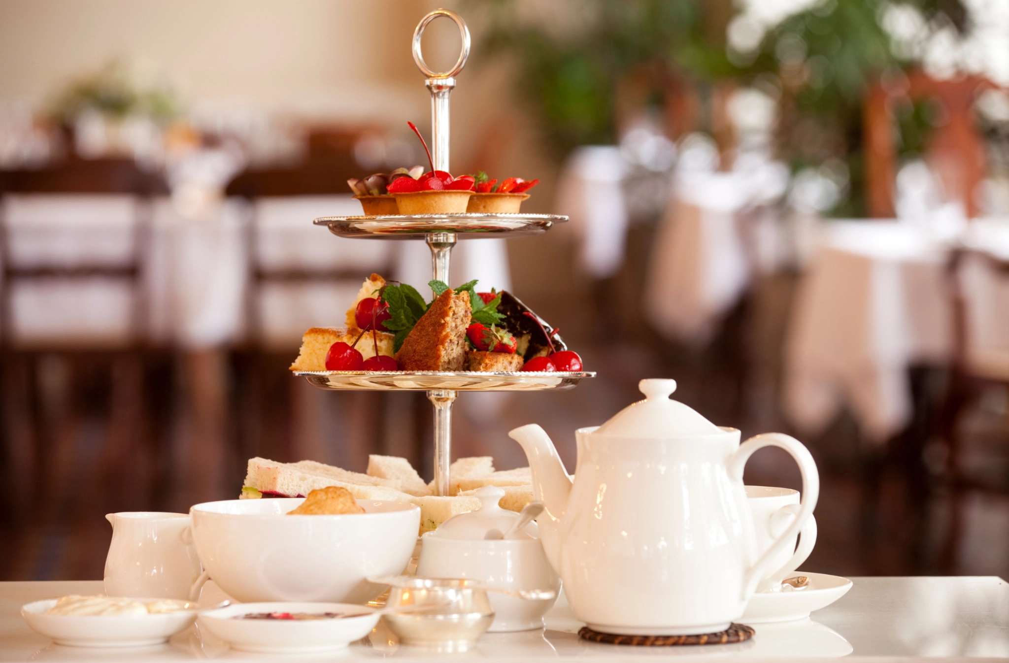 A white teapot and cups sit beside a tiered stand with pastries and sandwiches, set in a cozy, blurred café setting, evoking a warm, inviting atmosphere.