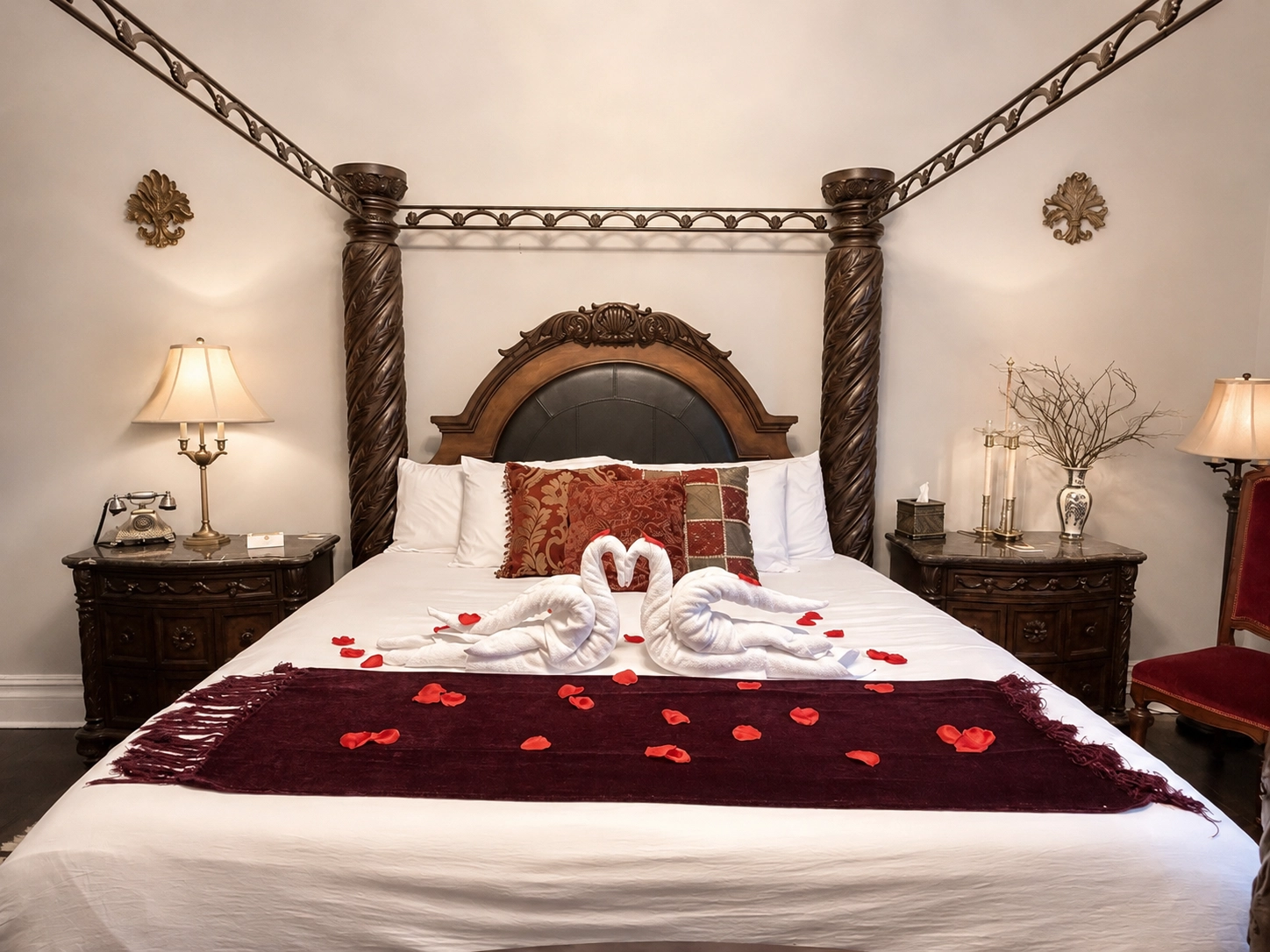 Luxurious bedroom with ornate wooden bed, white bedding, gold runner adorned with red rose petals. Warm lighting from elegant lamps creates a romantic ambiance.