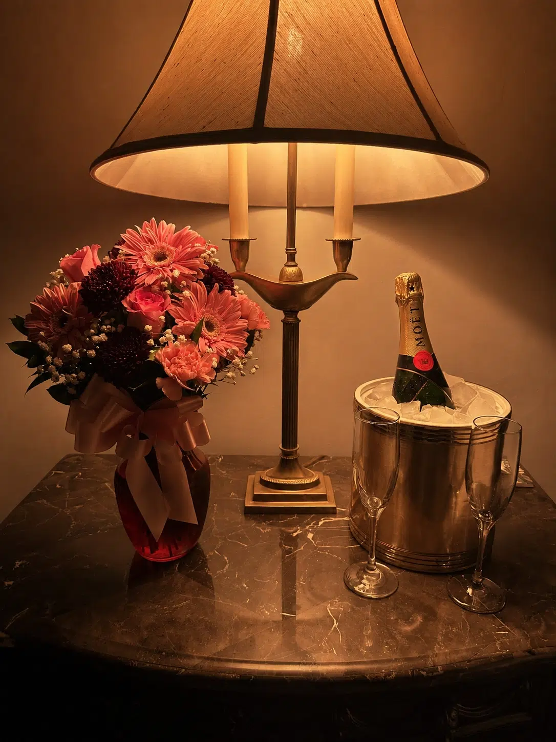 A warm-lit lamp, a bouquet of pink and maroon flowers in a vase, and a champagne bottle in an ice bucket with two flutes on a marble table.