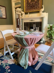 Elegant dining setting with a round table draped in pink and green fabric, surrounded by white chairs. The table holds glassware and fine china. A mantel with peacock sculptures and a decorative tapestry creates a sophisticated ambiance.