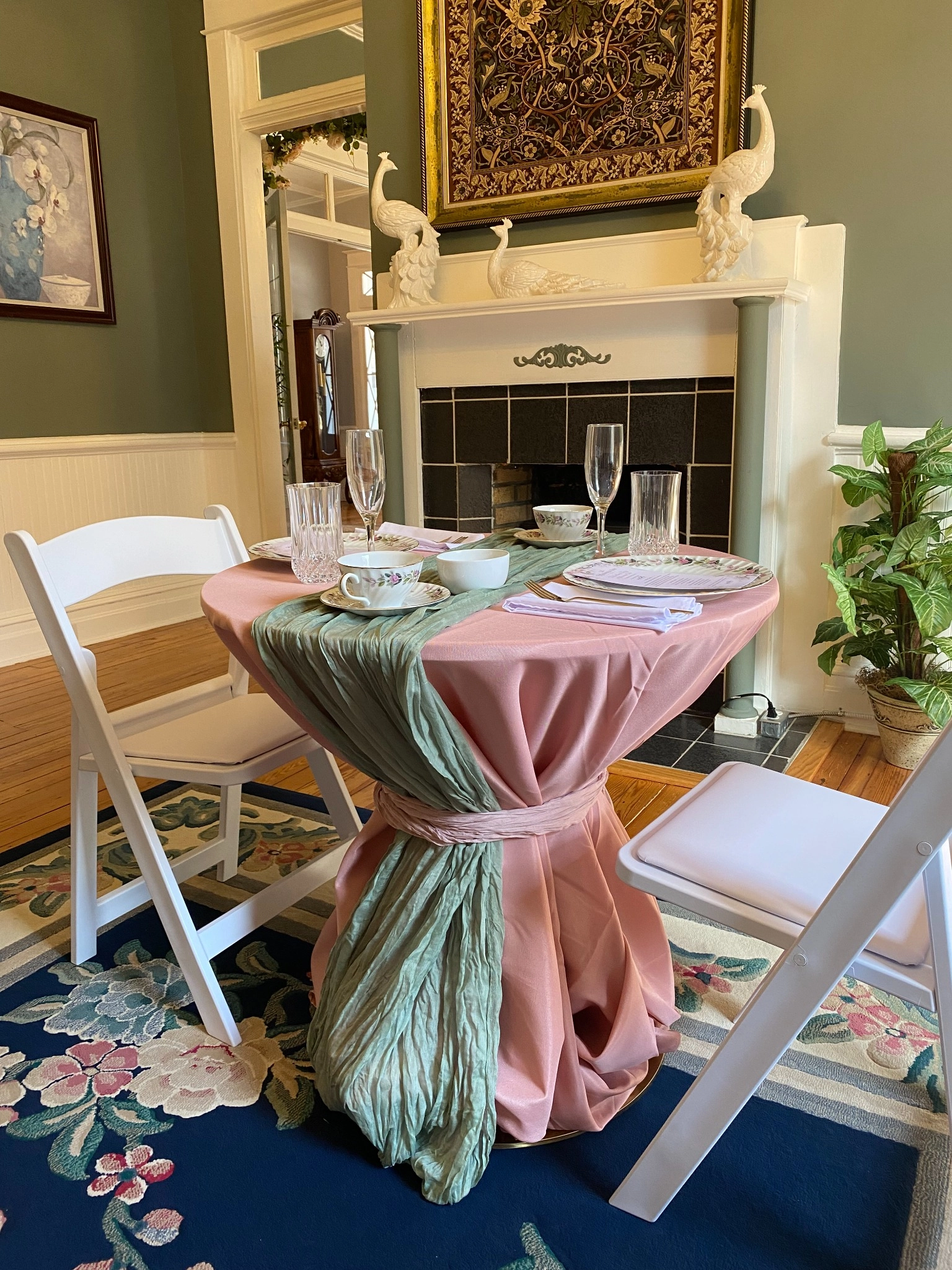 Elegant dining setting with a round table draped in pink and green fabric, surrounded by white chairs. The table holds glassware and fine china. A mantel with peacock sculptures and a decorative tapestry creates a sophisticated ambiance.