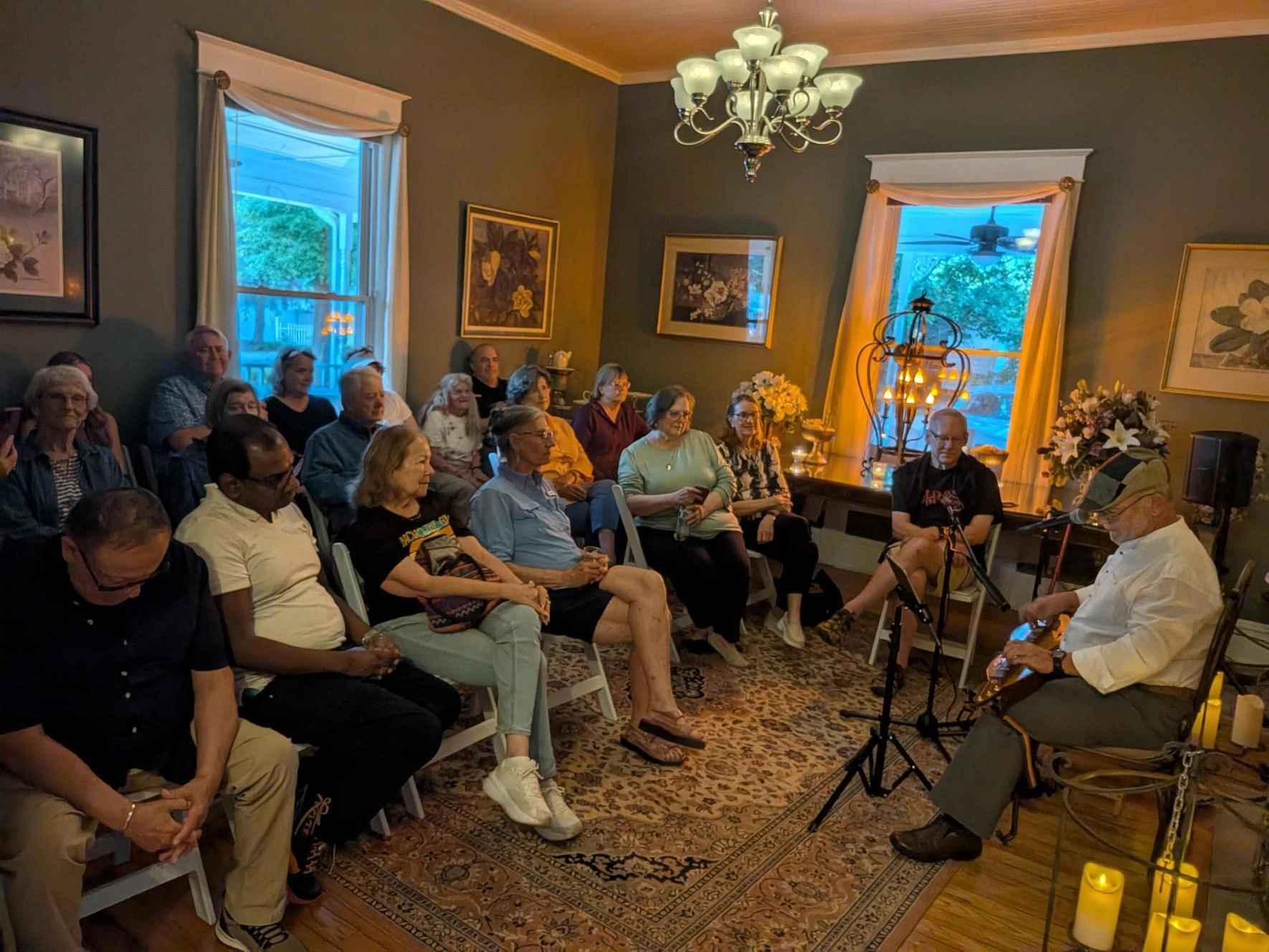 A cozy gathering in a warmly lit room shows an attentive audience seated on chairs. A man plays a stringed instrument. The scene is intimate and inviting.