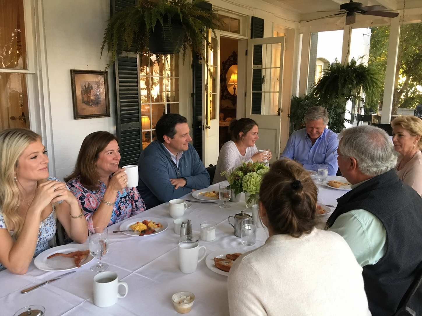 A group of eight people enjoy a casual meal on a veranda. They are smiling and engaged in conversation. The table is set with breakfast foods and coffee.