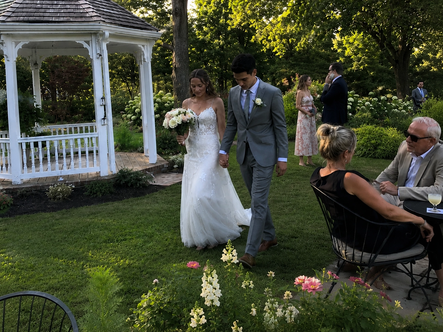 A bride in a white gown and groom in a gray suit walk hand in hand through a garden with guests seated nearby, next to a white gazebo.