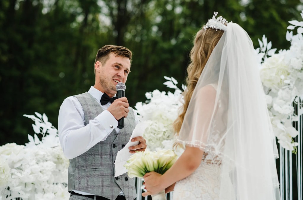 A groom in a grey vest holds a microphone, smiling while speaking to a bride in a white dress holding flowers. They stand before white floral decorations.
