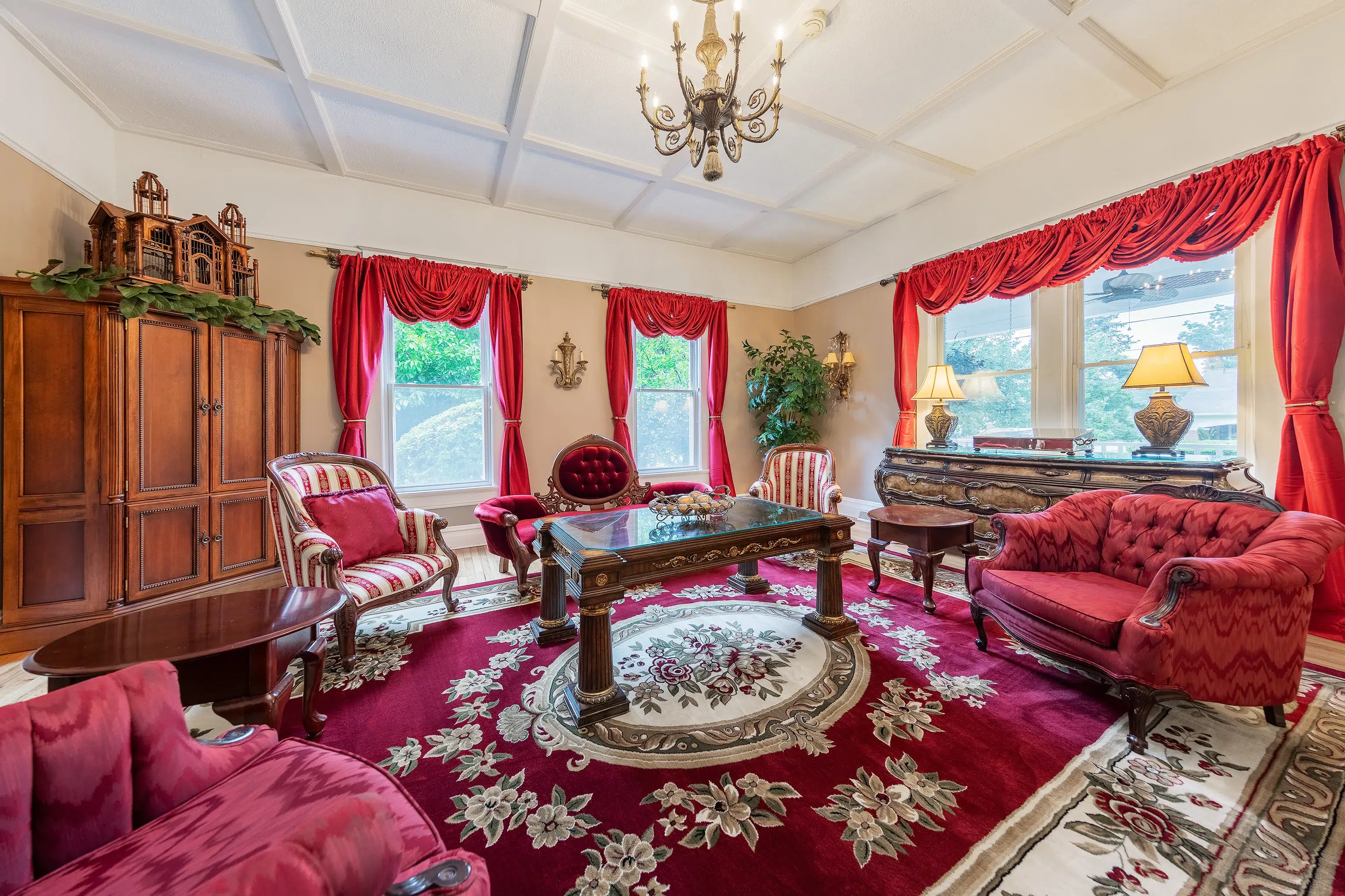 Spacious Victorian parlor with red drapes, antique seating, a floral rug, and a glass-top coffee table.