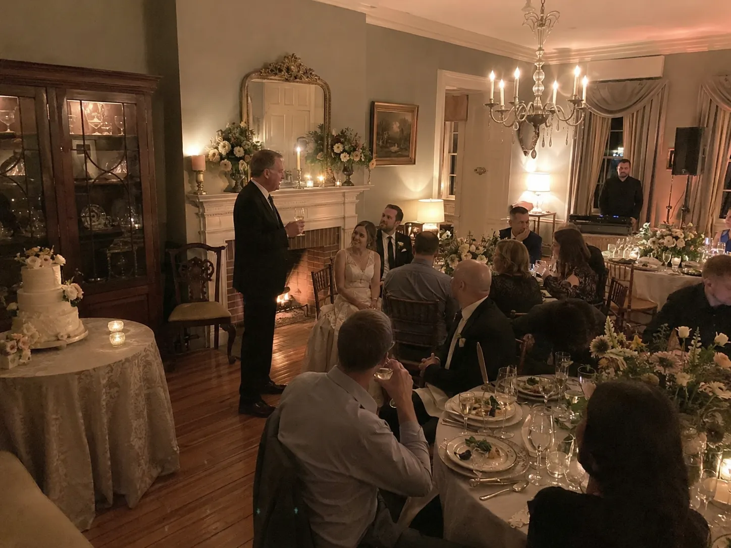 Elegant dinner gathering in a warmly lit room with people in formal attire seated around tables adorned with white tablecloths and floral centerpieces.