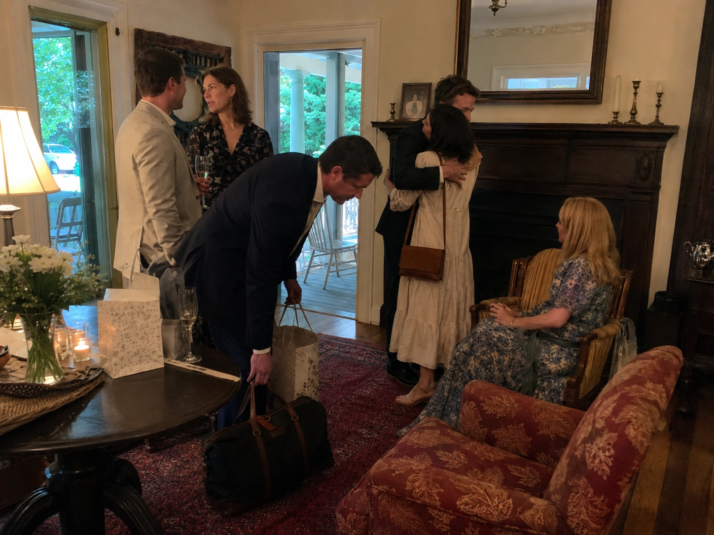 A cozy living room with people socializing. A couple hugs by a fireplace, another chats by a table with flowers and gifts, while a woman sits on a chair, observing.