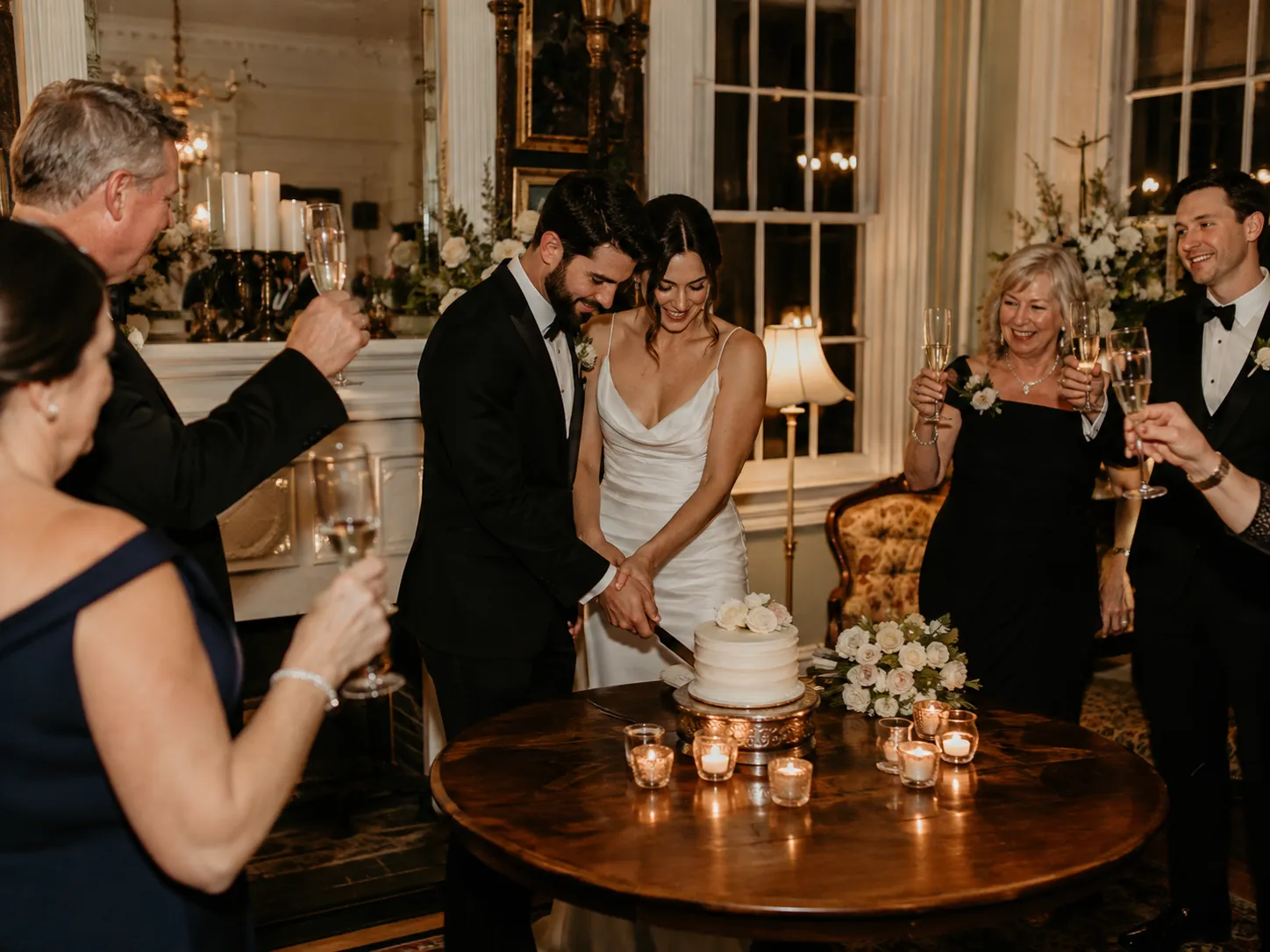 Bride and groom cutting their wedding cake as family and friends raise champagne glasses around them.